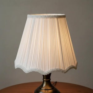 May include: A small, vintage-style table lamp with a pleated white lampshade. The shade has a scalloped edge with delicate trim. The base is a dark brass color. The lamp is on a wooden table.