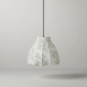 May include: A white pendant light fixture with a floral pattern in gray. The lampshade has a rounded, pleated shape and hangs from a black cord. The floral design is a delicate, botanical illustration.