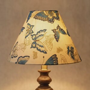 May include: A table lamp featuring a brown wooden base and a cream lampshade. The lampshade is adorned with a repeating pattern of blue butterflies. The lamp is illuminated, emitting a warm light.