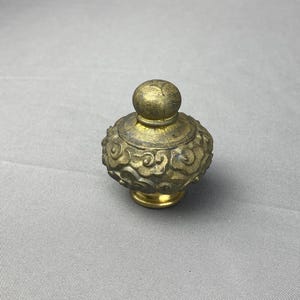 May include: A small, ornate, antique bottle stopper with a gold finish. The stopper features a rounded top and a detailed, cloud-like design around the body. The base is also gold, creating a cohesive, vintage aesthetic.