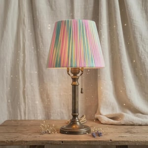 Puede incluir: Una lámpara de mesa de estilo vintage con una pantalla a rayas multicolores. La pantalla presenta rayas verticales en rosa, amarillo, azul y verde. La lámpara tiene una base y un vástago de latón, y una cadena. La lámpara está sobre una mesa de madera.