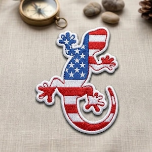 May include: An embroidered patch in the shape of a lizard, featuring a design of the American flag with a blue field of white stars on the upper body and red and white horizontal stripes covering the remainder of the lizard's body and tail.