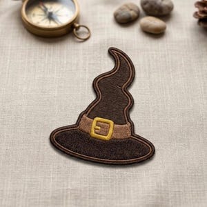 May include: Embroidered patch of a brown witch hat with a wavy top and a tan band featuring a gold buckle. The patch is likely for clothing or accessories, perfect for Halloween or a costume.