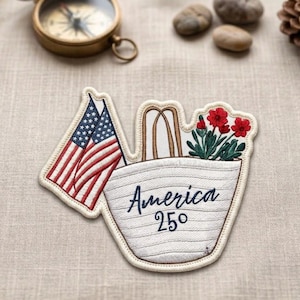 May include: Embroidered patch featuring two American flags and a tote bag filled with red flowers. The tote bag has the word "America" and the number "250" embroidered on it. The patch is cream-colored with a stitched border.