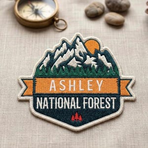 May include: Embroidered patch featuring a mountain range with a rising sun. The patch has the text "ASHLEY NATIONAL FOREST" in orange and white. The patch is bordered in white and has a navy blue background.