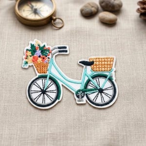 May include: Embroidered patch of a light blue bicycle with a basket of colorful flowers in the front and a basket in the back. The bicycle has black wheels with white spokes and a black seat. The patch is outlined in white.