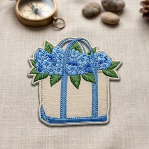 May include: Embroidered patch featuring a tote bag design filled with blue hydrangea flowers and green leaves. The bag has blue handles and trim, set against a beige background. The patch is likely for clothing or accessories.