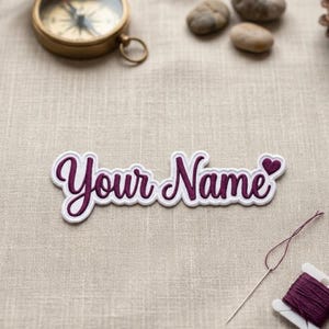 May include: A custom embroidered patch featuring the dark purple script text Your Name with a small heart icon at the end. The patch has a white border and is displayed alongside a brass compass, smooth stones, and a spool of purple thread.