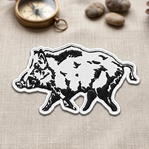 May include: A white patch with a black outline of a wild boar. The patch is in the shape of the animal, with details of the boar's fur and features. A vintage compass and small stones are in the background.