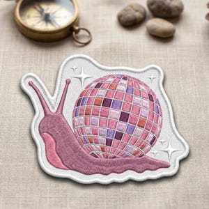May include: Embroidered patch featuring a pink snail with a disco ball shell. The snail is a light pink color with a white border and star accents. The disco ball is made up of pink, purple, and white squares.