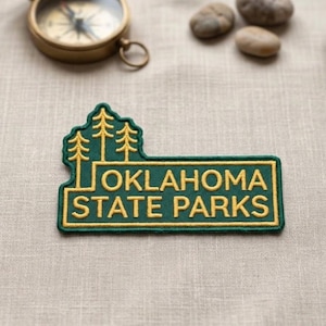 May include: A green and gold embroidered patch with the text "OKLAHOMA STATE PARKS" and a tree design. The patch is rectangular with a tree-shaped top. A vintage compass and small stones are in the background.