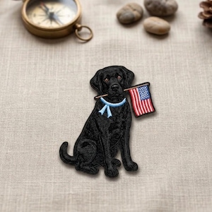 May include: Embroidered patch featuring a black Labrador Retriever sitting upright, holding a small American flag in its mouth. The dog wears a light blue ribbon around its neck. The flag has red and white stripes and a blue field with white stars.