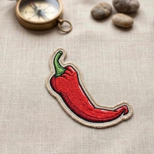 May include: Embroidered red chili pepper patch with a green stem and tan border. The patch is on a tan fabric surface. A vintage brass compass and small stones are in the background.