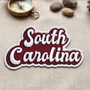May include: Embroidered patch with the words "South Carolina" in white script on a maroon background. The patch has a white and maroon border. A vintage compass and small stones are in the background.