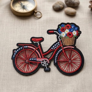 May include: Embroidered patch of a red bicycle with a basket of flowers. The bicycle has red wheels and a black outline. The basket contains red, white, and blue flowers. The design includes stars and stripes.