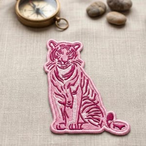 May include: A pink embroidered patch featuring a detailed tiger design. The tiger is depicted in a seated position with intricate stripes and facial features. The patch is likely for clothing or accessories.