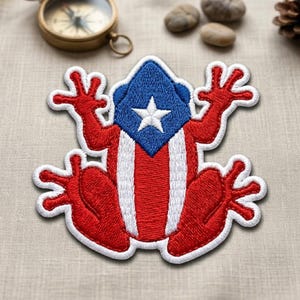 May include: Embroidered patch featuring a frog design in the colors of the Puerto Rican flag. The frog has a blue head with a white star, red arms and legs, and red and white stripes on its body. The patch has a white border.