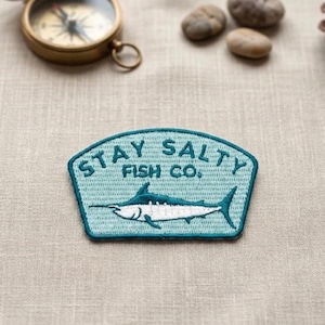 May include: A turquoise patch with the text "STAY SALTY FISH CO." and a white marlin illustration. The patch is bordered in a darker turquoise and is set against a neutral background. A vintage brass compass is in the background.