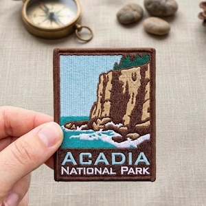 May include: Embroidered patch featuring a coastal landscape with the text "ACADIA NATIONAL PARK." The patch has a brown border, a light blue sky, and a turquoise ocean. The design includes a cliffside and waves.