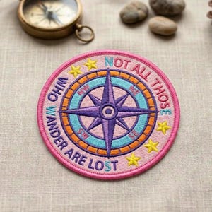 May include: A round, embroidered patch with a compass rose design. The patch features a pink border with the text "NOT ALL THOSE WHO WANDER ARE LOST." The compass rose is purple, blue, and orange, with yellow star accents.