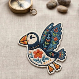 May include: Embroidered patch featuring a cartoon puffin bird with a white face, orange beak, and blue wings decorated with orange and green floral designs. The patch is on a neutral linen surface, with a brass compass and small stones in the background.