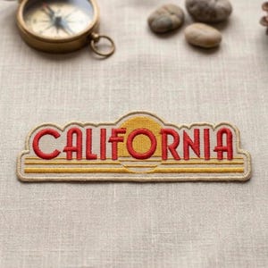 May include: Embroidered patch with the word "CALIFORNIA" in red, with a yellow sun and horizontal lines. The patch is beige with a gold border. A vintage compass and small stones are in the background.