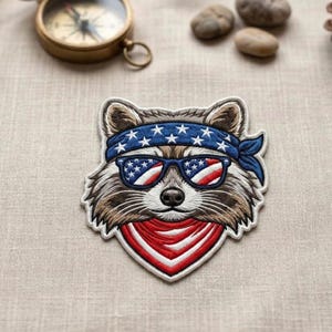 May include: Embroidered patch featuring a raccoon wearing sunglasses with a red, white, and blue American flag design. The raccoon has a blue bandana with white stars and a red and white bandana around its neck. The patch is on a neutral linen surface.