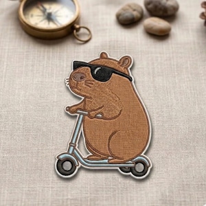 May include: Embroidered patch featuring a brown capybara wearing sunglasses and riding a light blue scooter. The patch has a detailed design and a black border. The background is a neutral beige fabric.