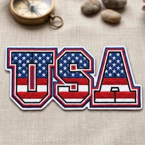 May include: A patriotic USA patch with a bold design. The letters are filled with a blue field of white stars and red and white stripes, outlined in blue and white. The patch is likely for clothing or accessories.