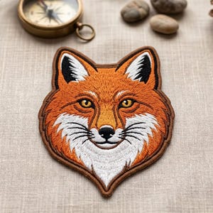 May include: Embroidered patch featuring a detailed fox head with orange, white, and black fur. The patch has a brown border and is designed with intricate stitching. The fox has yellow eyes and a black nose.