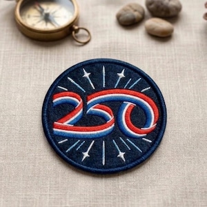May include: A round, navy blue embroidered patch with the number "250" in red, white, and blue. The design includes radiating lines and stars, with a navy blue border. A vintage brass compass and small stones are in the background.