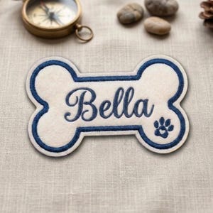 May include: A bone-shaped patch with an off-white felt base and a dark blue embroidered border. The name Bella is embroidered in dark blue script in the center, accompanied by a small dark blue paw print icon on the right side of the patch.