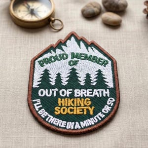 May include: Embroidered patch with the text "PROUD MEMBER OF OUT OF BREATH HIKING SOCIETY I'LL BE THERE IN A MINUTE OR SO." The patch features a mountain and tree design in green, white, and brown. A compass and small stones are in the background.