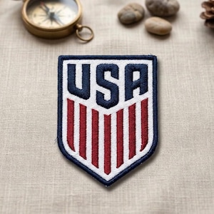 May include: Embroidered patch with the word "USA" in navy blue letters. The patch has a white border and a shield design with red and white vertical stripes. The patch is on a beige fabric surface.