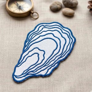 May include: An embroidered patch in the shape of an oyster shell. The patch features a white interior with blue outline and detailing. The patch is on a neutral-colored fabric surface.