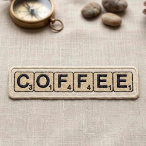 May include: A beige embroidered patch spelling "COFFEE" with black letters, resembling Scrabble tiles. The patch is rectangular with a stitched border. A vintage brass compass and small stones are in the background.
