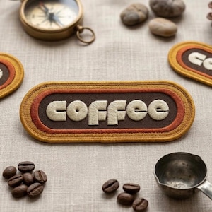 May include: An embroidered patch with the word "coffee" in cream-colored thread, set against a brown background with orange and gold borders. Coffee beans and a small metal scoop are also visible, suggesting a coffee theme.