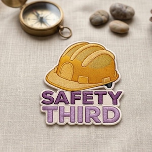 May include: Embroidered patch featuring a yellow construction helmet with the text "SAFETY THIRD" in purple. A vintage brass compass and small stones are in the background. The patch is likely for a jacket or bag.