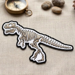 May include: A dinosaur skeleton patch with a black border. The patch features a detailed illustration of a Tyrannosaurus Rex skeleton in shades of beige and white. The patch is designed for sewing or ironing onto fabric.