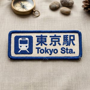 May include: Embroidered patch with a blue border and a cream-colored center. The patch features a train icon and the text "東京駅 Tokyo Sta." in blue. A compass, small stones, and a pinecone are in the background.