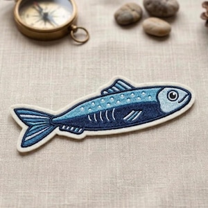 May include: Embroidered patch in the shape of a fish, featuring shades of blue and white. The fish has a detailed design with scales, fins, and an eye. The patch is likely for clothing or accessories.