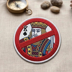 May include: Embroidered patch featuring a king card with a red prohibition symbol. The king is depicted in a crown and royal attire, with a spade symbol and the letter "K". The patch has a white border and is circular.