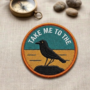 May include: Embroidered patch with a black crow design, perched on a rock, set against a teal and yellow background. The patch features the text "TAKE ME TO THE" in white. A vintage compass and small stones are in the background.