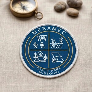 May include: A round, blue Meramec State Park patch with white stitching. The patch features a gold border and four quadrants with white illustrations of outdoor activities and the text "MERAMEC STATE PARK MISSOURI". A vintage compass and small stones are in the background.