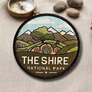 May include: Embroidered patch featuring a landscape scene with rolling green hills, mountains, and hobbit-hole homes. The patch has the text "THE SHIRE NATIONAL PARK" in white lettering. The patch is round with a black border.