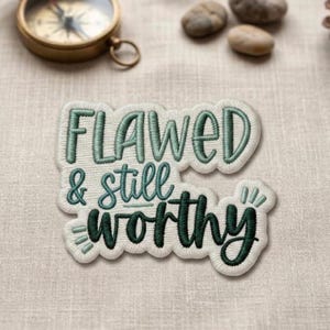 May include: An embroidered patch with the words "FLAWED & still WORTHY" in teal thread on a white background. The patch has a decorative border. A vintage compass and small stones are in the background.