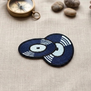 May include: Two navy blue embroidered patches in the shape of vinyl records. Each patch has a white and light blue design. The patches are on a beige linen surface, with a brass compass and small stones in the background.