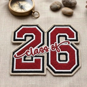 May include: A red and white embroidered patch with the number "26" and the words "Class of" in cursive. The patch has a black border and a tan background. A vintage compass and small stones are in the background.