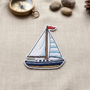 May include: An embroidered fabric patch shaped like a sailboat. The boat has a dark blue hull with a gold trim, a brown mast, and white sails. One sail features light blue horizontal stripes, and a small red flag is attached to the top of the mast.