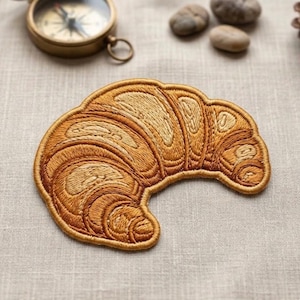 May include: Embroidered patch in the shape of a croissant, featuring detailed stitching in shades of brown and tan. The patch is designed to resemble the texture of a freshly baked pastry. A compass and small stones are in the background.
